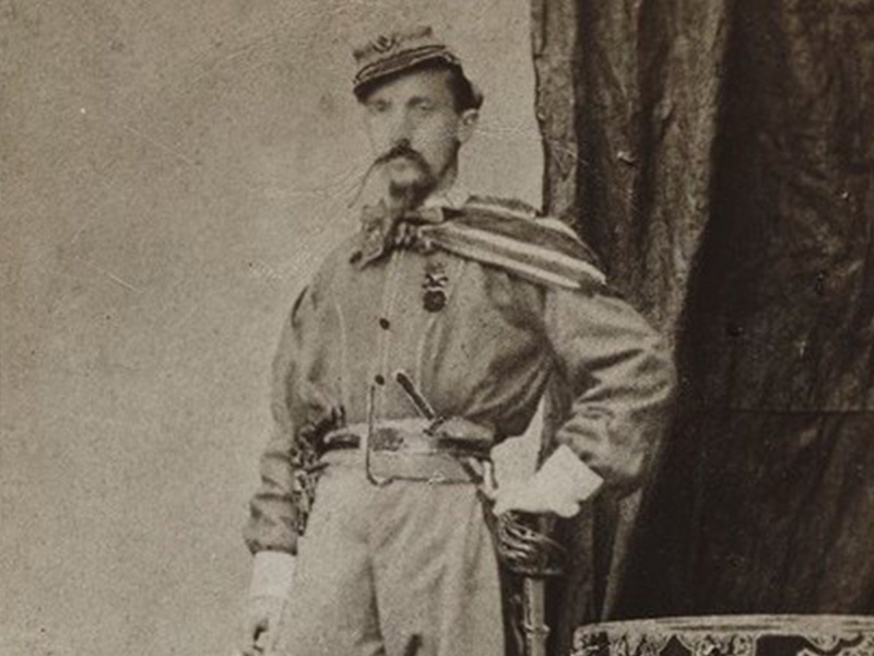 Foto di figura intera, ritratto in piedi, in divisa garibaldina. Sullo sfondo un drappo, a sinistra un piccolo tavolo rotondo